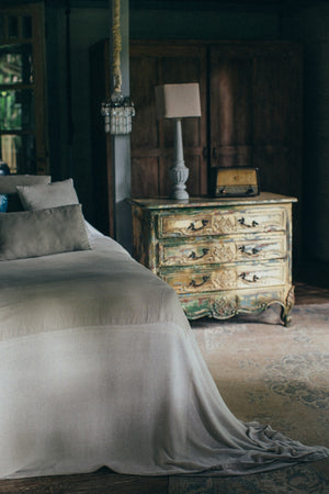 Corner of a bed and a bedside chest with a lamp in a dark bedroom