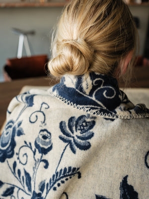 A person viewed from behind covered by a cream-navy colored blanket with a navy blue floral and scroll pattern, styled with a low bun.