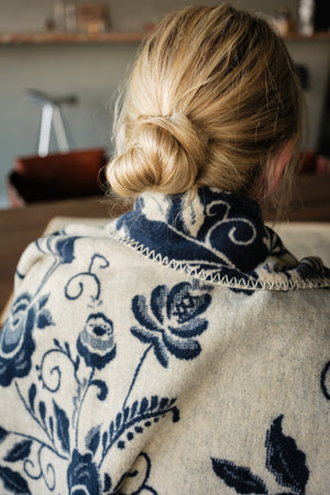 A person viewed from behind covered by a cream-navy colored blanket with a navy blue floral and scroll pattern, styled with a low bun.